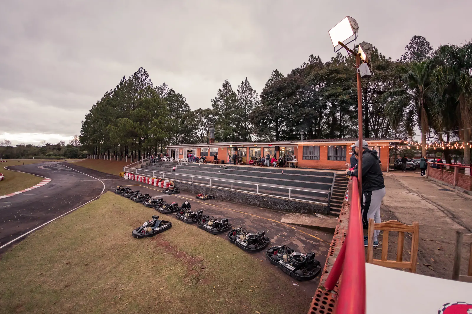 Karts de aluguel alinhados na grama ao lado da pista, diante da varanda de tijolo com arquibancada e varal de lâmpadas do kartódromo